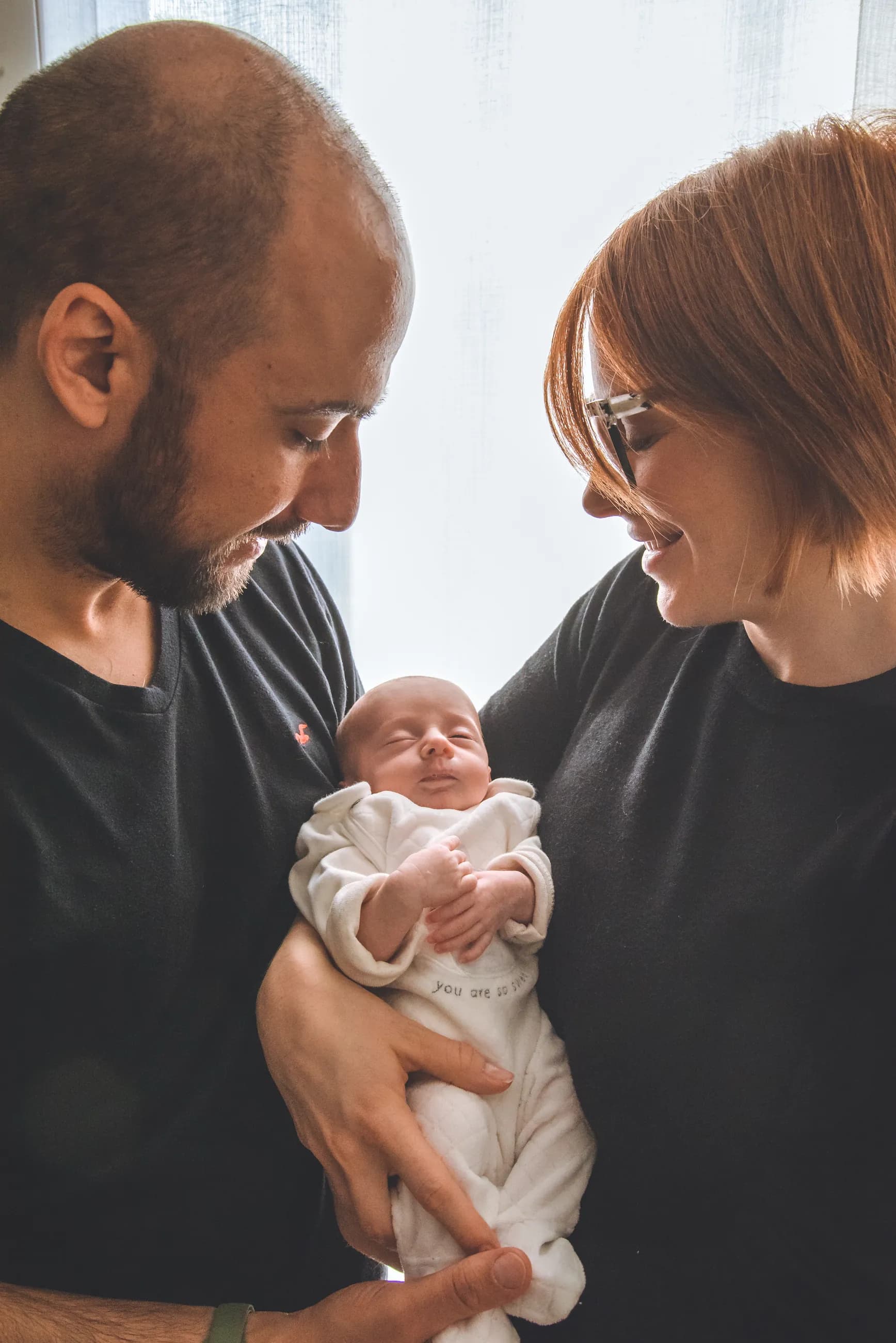 Mamma e papà, di profilo contro la luce di una finestra, guardano il loro neonato in braccio