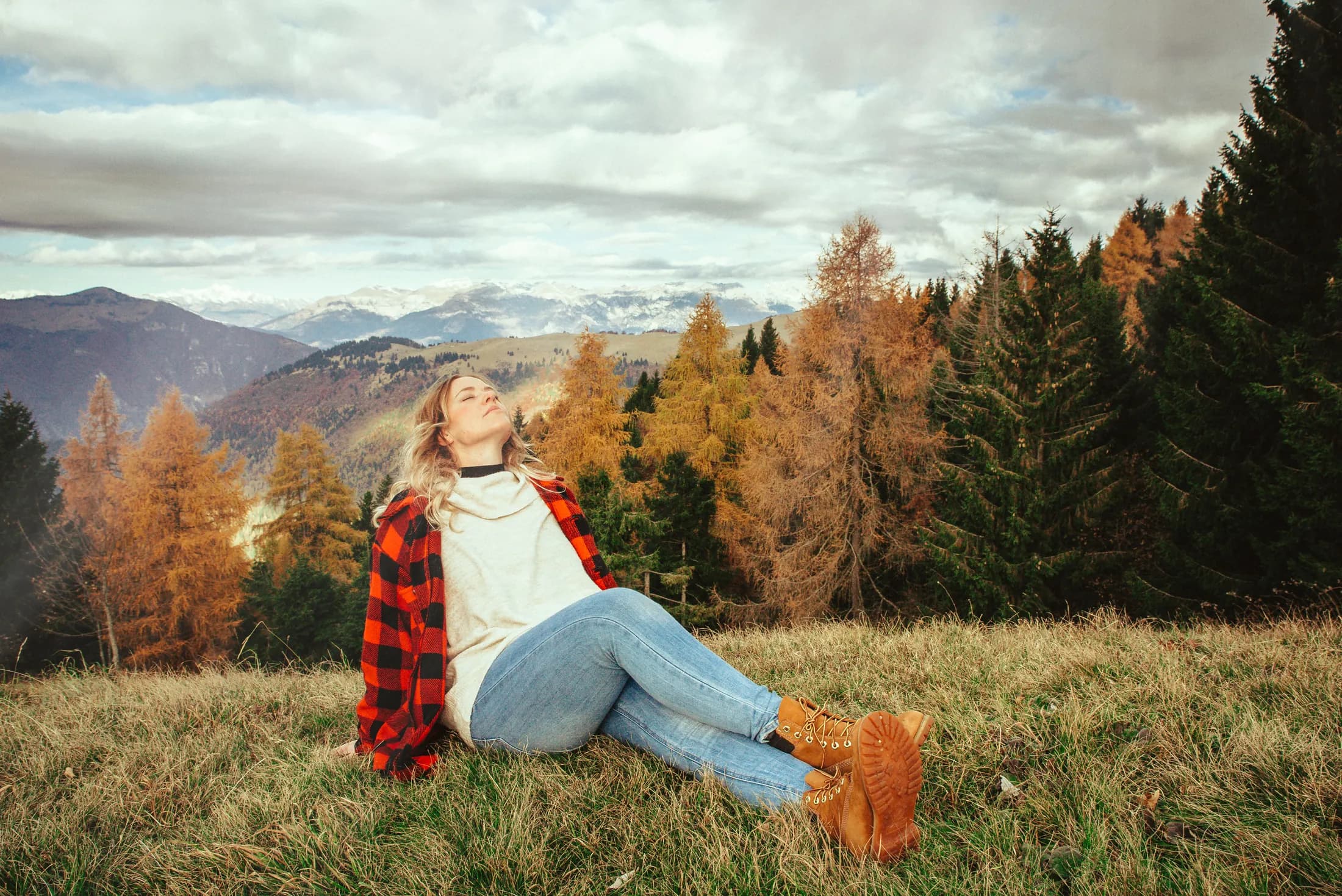 Una donna seduta su un prato di montagna alza il viso verso la luce, tra i larici d’autunno